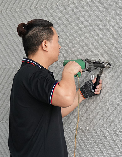Ein Handwerker in schwarzem shirt arbeitet seitlich stehend mit einer Tuftingpistole an einem grauen Teppich mit diagonalen Linien