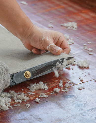 Eine Hand führt eine große Schere entlang der Kante eines Teppichs, schneidet überschüssiges Material ab, Teppichfasern liegen auf der roten Arbeitsfläche
