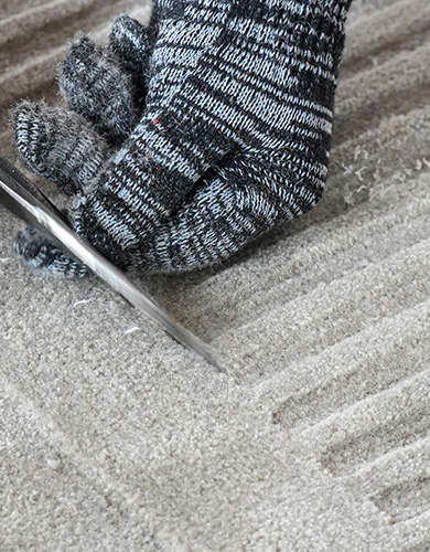 Nahaufnahme einer Hand mit grau-weiß gestreiftem Handschuh beim Texturschneiden eines beigen Teppichs