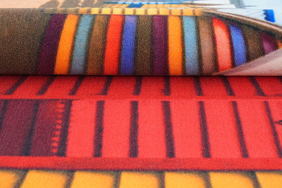 Ein maßgefertigter bedruckter Nylon-Teppich beim Aufrollen, der die blaue Oberseite und einen Teil des darunterliegenden Designs mit blauen, orangefarbenen und dunklen Tönen zeigt und die Textur seiner geschnittenen Flore präsentiert.
